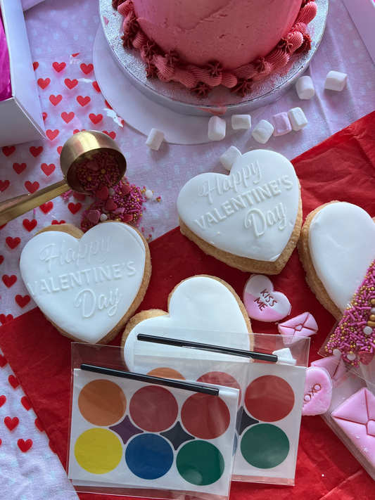 Heart-shaped cookies with 'Happy Valentine's Day' text on a table with a watercolor palette and pink cake.